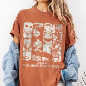 May include: A rust-colored short-sleeve t-shirt featuring a distressed white graphic print of characters from Tim Burton's The Nightmare Before Christmas, including Jack Skellington, Sally, and Oogie Boogie, arranged in four vertical panels.