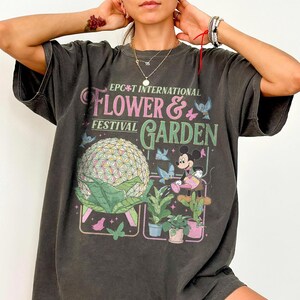 May include: Dark gray t-shirt featuring the text "EPCOT International Flower & Garden Festival". The graphic includes a geometric sphere, Mickey Mouse watering plants, and butterflies. The shirt has short sleeves and a relaxed fit.