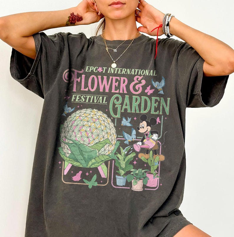 May include: Dark gray t-shirt featuring the text "EPCOT International Flower & Garden Festival". The graphic includes a geometric sphere, Mickey Mouse watering plants, and butterflies. The shirt has short sleeves and a relaxed fit.