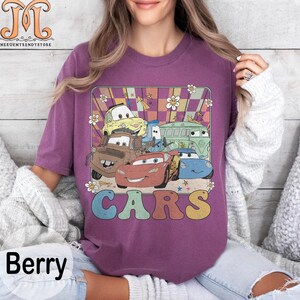 May include: A purple t-shirt with a retro design featuring a group of cartoon cars from the movie Cars. The cars are in a colorful checkered pattern with the word "CARS" in a rainbow font.