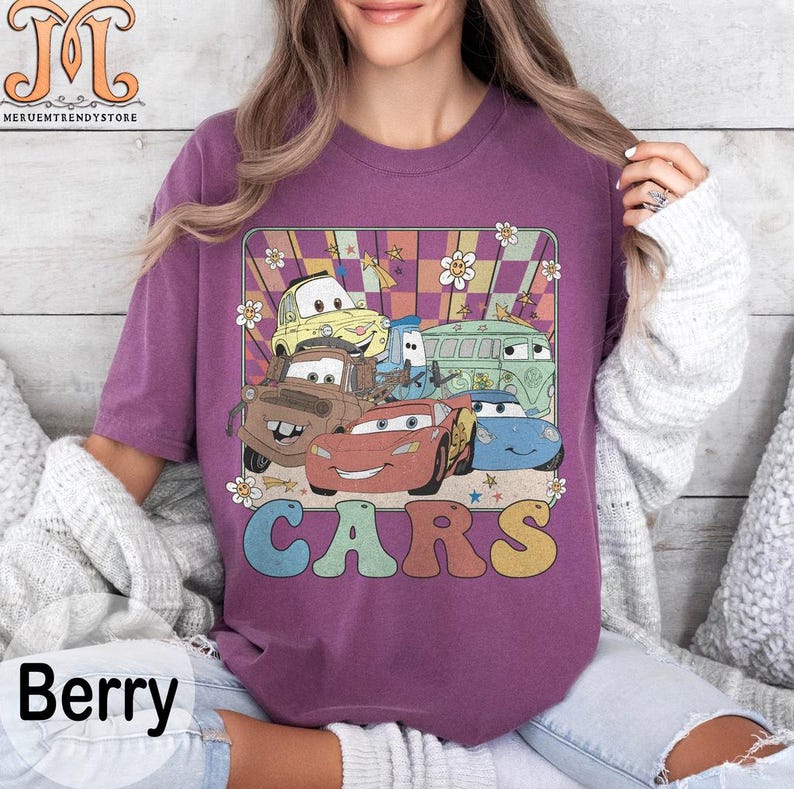 May include: A purple t-shirt with a retro design featuring a group of cartoon cars from the movie Cars. The cars are in a colorful checkered pattern with the word "CARS" in a rainbow font.