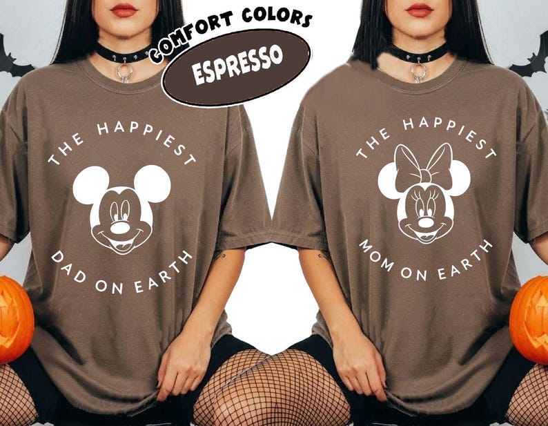 May include: Two espresso-colored t-shirts with white graphic designs. One shirt features Mickey Mouse with the text "The Happiest Dad on Earth," and the other features Minnie Mouse with "The Happiest Mom on Earth." The shirts have a comfort colors label.