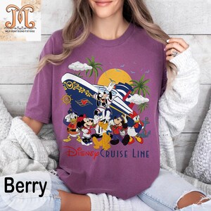 May include: Berry-colored t-shirt featuring a Disney Cruise Line graphic. The design includes Mickey Mouse, Minnie Mouse, Donald Duck, and Goofy, along with a cruise ship, palm trees, and a sunset. The text "Disney Cruise Line" is printed below the characters.