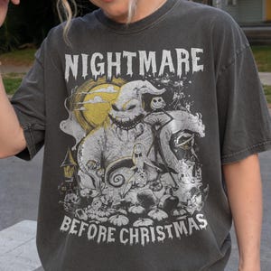 May include: A charcoal gray short-sleeve t-shirt featuring a graphic print of characters from The Nightmare Before Christmas. The design includes Oogie Boogie, Jack Skellington, and Sally, with the text NIGHTMARE BEFORE CHRISTMAS printed.