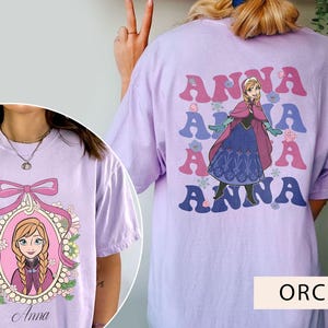 May include: Two purple t-shirts with different designs featuring the character Anna from the movie Frozen. One shirt has a graphic of Anna with the word "Anna" repeated five times. The other shirt has a graphic of Anna in a frame with the word "Anna" below the frame.