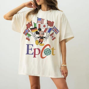 Könnte beinhalten: Cremefarbenes T-Shirt mit einem Aufdruck von Mickey Mouse, der Flaggen aus verschiedenen Ländern hält. Das Wort "Epcot" ist in bunten Buchstaben unter dem Aufdruck gedruckt, mit einem Globus, der das "o" ersetzt.