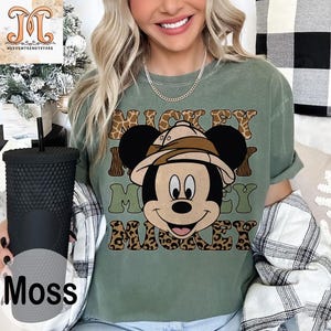 May include: A green t-shirt with a cartoon image of Mickey Mouse wearing a safari hat. The text "Mickey Mouse" is printed on the shirt in a brown, green, and leopard print pattern.