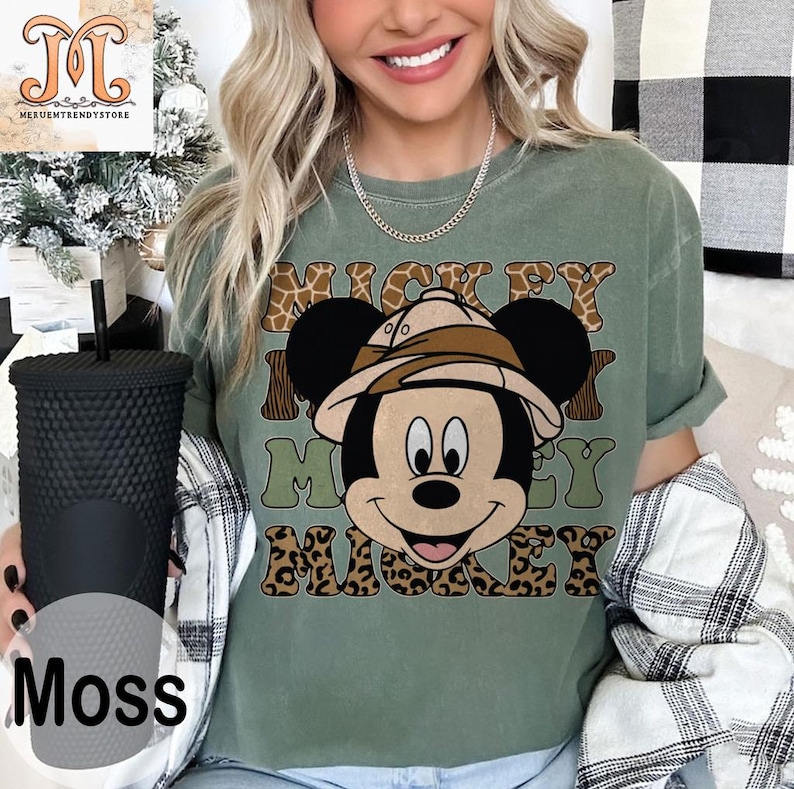 May include: A green t-shirt with a cartoon image of Mickey Mouse wearing a safari hat. The text "Mickey Mouse" is printed on the shirt in a brown, green, and leopard print pattern.
