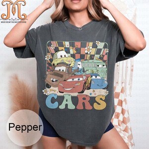 May include: A dark gray t-shirt with a retro-style graphic of cartoon cars from the movie Cars. The graphic features a checkered background with stars and flowers. The text "CARS" is printed in rainbow colors below the graphic.
