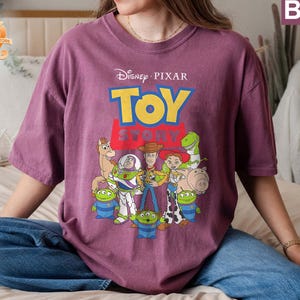 May include: Berry-colored t-shirt featuring the Disney Pixar Toy Story logo and characters. The design includes Woody, Buzz Lightyear, and other characters from the animated film. The shirt is a crew neck and has short sleeves.