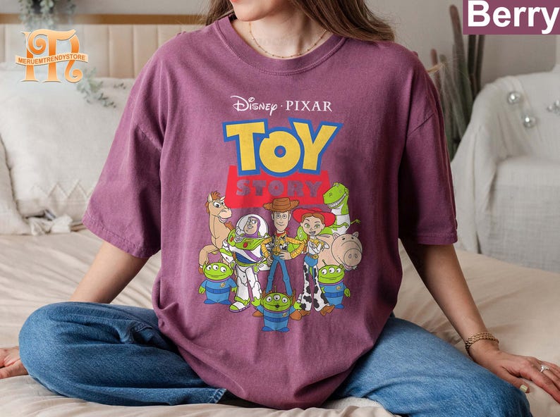 May include: Berry-colored t-shirt featuring the Disney Pixar Toy Story logo and characters. The design includes Woody, Buzz Lightyear, and other characters from the animated film. The shirt is a crew neck and has short sleeves.