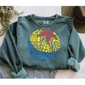 May include: A teal crewneck sweatshirt featuring a colorful graphic. The graphic is a circle divided into three sections: a red star, yellow figures, and blue figures. The sweatshirt has a relaxed fit and rolled cuffs.