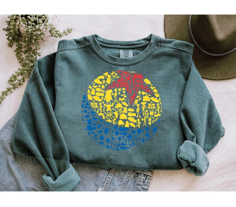 May include: A teal crewneck sweatshirt featuring a colorful graphic. The graphic is a circle divided into three sections: a red star, yellow figures, and blue figures. The sweatshirt has a relaxed fit and rolled cuffs.