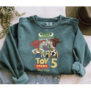 May include: Teal green sweatshirt featuring a graphic of characters from Toy Story 5. The design includes Woody, Buzz Lightyear, Jessie, and other characters. The sweatshirt has a crew neck and long sleeves.