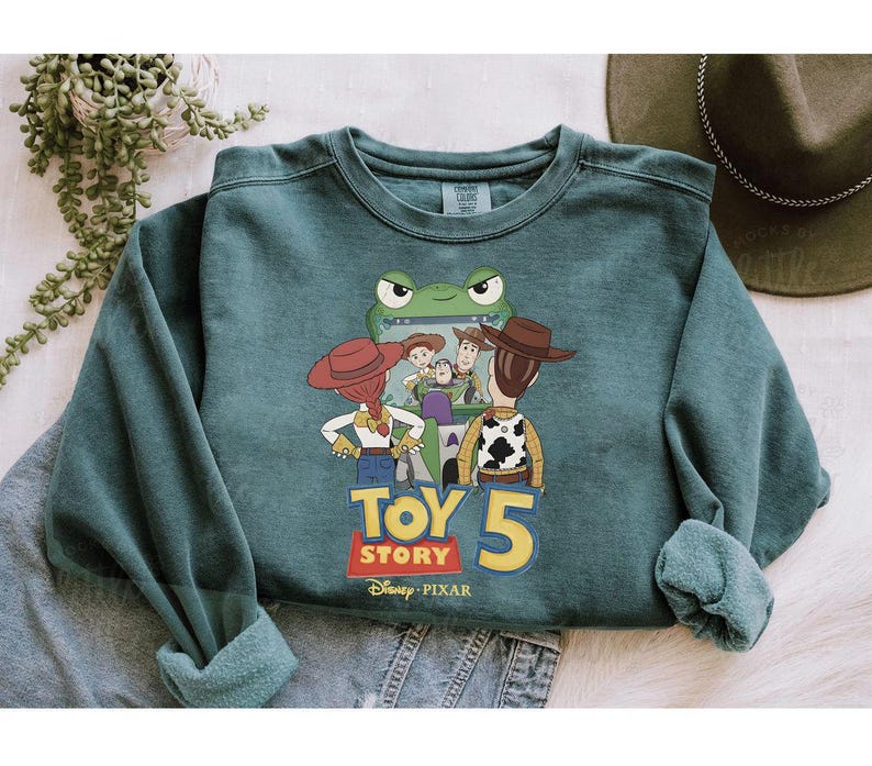 May include: Teal green sweatshirt featuring a graphic of characters from Toy Story 5. The design includes Woody, Buzz Lightyear, Jessie, and other characters. The sweatshirt has a crew neck and long sleeves.