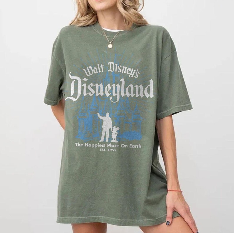 Puede incluir: Camiseta verde oliva con el texto blanco "Walt Disney's Disneyland", con una silueta azul de una persona y Mickey Mouse. El texto "The Happiest Place On Earth" y "EST. 1955" est&aacute;n debajo.