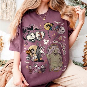 May include: A dark purple t-shirt featuring a graphic print of characters and motifs from The Nightmare Before Christmas, including Jack Skellington, Sally, Oogie Boogie, Zero, and various Halloween-themed icons like bats, pumpkins, and spiderwebs.