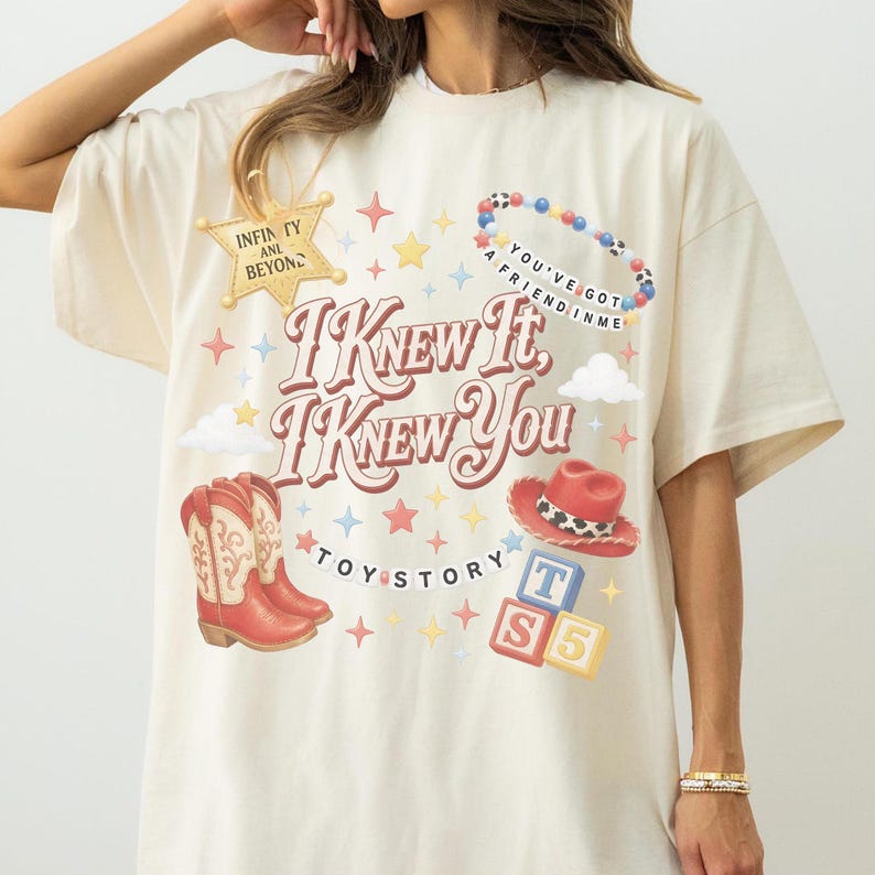 May include: A cream-coloured t-shirt featuring a graphic print with the text I KNEW IT, I KNEW YOU, TOY STORY, INFINITY AND BEYOND, and YOU'VE GOT A FRIEND IN ME. The design includes a sheriff badge, a beaded bracelet, cowboy boots, a hat, and blocks.