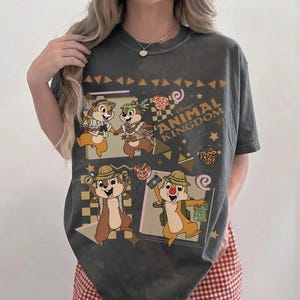 May include: A charcoal gray short-sleeve t-shirt featuring a graphic print of Chip and Dale in safari gear. The design includes the text Disney ANIMAL KINGDOM, along with various geometric shapes, stars, and small animal-print accents.