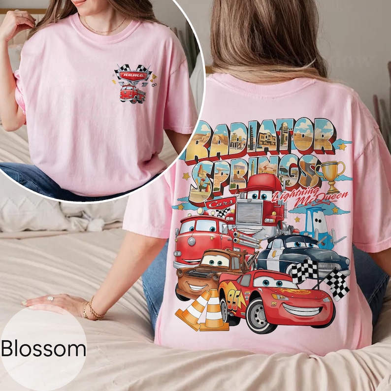 Puede incluir: Camiseta rosa claro con un gr&aacute;fico colorido de personajes de Cars y la inscripci&oacute;n "Radiator Springs" en la espalda. La parte delantera tiene un gr&aacute;fico m&aacute;s peque&ntilde;o con un coche y banderas a cuadros. La palabra "Blossom" est&aacute; en la parte inferior.