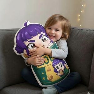 Może przedstawiać: Pluszowa poduszka w kształcie postaci z kreskówki z fioletowymi włosami i dużymi oczami. Poduszka jest zielono-żółta z obrazkami jedzenia. Dziecko trzyma poduszkę.