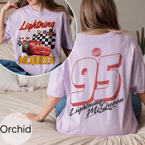 May include: Orchid-colored t-shirt featuring Lightning McQueen from the Cars movie. The front graphic includes the text "Lightning McQueen" and "95" with a checkered flag. The back displays a large "95" and "Lightning McQueen" in red and black.