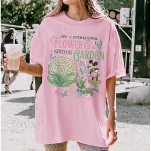 May include: A light pink t-shirt featuring the text "EPCOT INTERNATIONAL FLOWER & FESTIVAL GARDEN" with a graphic of Spaceship Earth, Mickey Mouse, and plants. The design includes butterflies and other decorative elements.