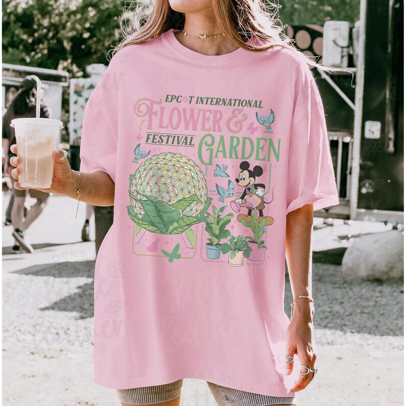 May include: A light pink t-shirt featuring the text "EPCOT INTERNATIONAL FLOWER & FESTIVAL GARDEN" with a graphic of Spaceship Earth, Mickey Mouse, and plants. The design includes butterflies and other decorative elements.