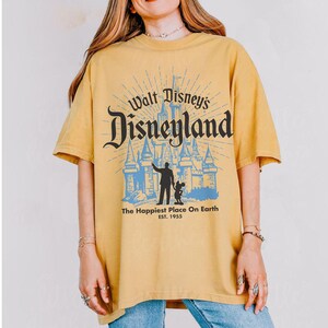 Puede incluir: Camiseta de color amarillo mostaza con el texto "Walt Disney's Disneyland" en una fuente estilizada, un gr&aacute;fico de castillo azul y la frase "The Happiest Place On Earth". La camiseta tiene mangas cortas y cuello redondo.