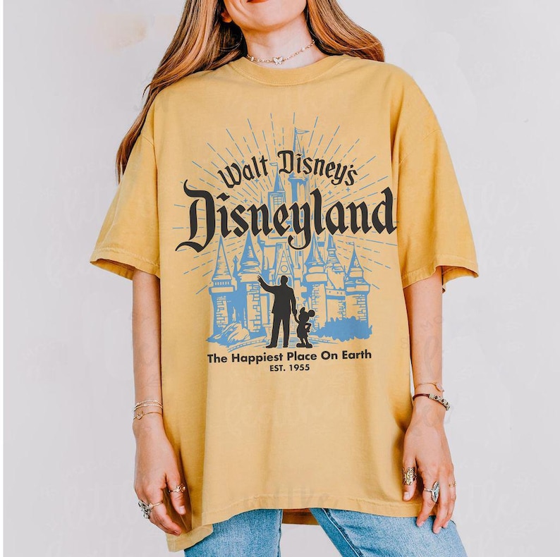 Puede incluir: Camiseta de color amarillo mostaza con el texto "Walt Disney's Disneyland" en una fuente estilizada, un gr&aacute;fico de castillo azul y la frase "The Happiest Place On Earth". La camiseta tiene mangas cortas y cuello redondo.