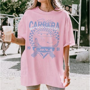 May include: Pink t-shirt featuring a light blue graphic of a cartoon car with the text "Sally Carrera" and "Cars 1951". The design includes checkered flags and a circular emblem. The shirt has a relaxed fit.