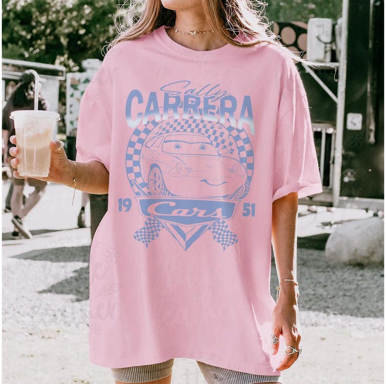 May include: Pink t-shirt featuring a light blue graphic of a cartoon car with the text "Sally Carrera" and "Cars 1951". The design includes checkered flags and a circular emblem. The shirt has a relaxed fit.