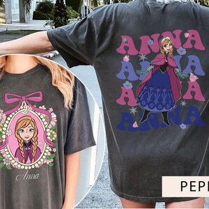 May include: Two gray t-shirts with a graphic print of Anna from Frozen. One shirt features a cartoon image of Anna in a frame with flowers and the text "Anna". The other shirt features a cartoon image of Anna in a blue and purple dress with the text "Anna" repeated in a retro font.