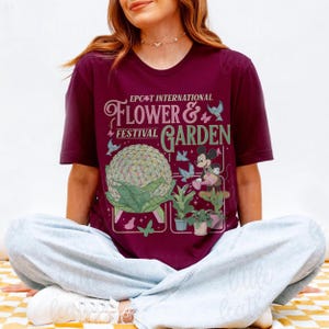 May include: A maroon t-shirt featuring the text "EPCOT International Flower & Garden Festival" with a graphic of Spaceship Earth, Mickey Mouse, and plants. The design includes butterflies and birds.
