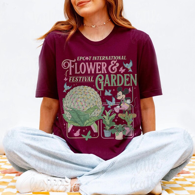May include: A maroon t-shirt featuring the text "EPCOT International Flower & Garden Festival" with a graphic of Spaceship Earth, Mickey Mouse, and plants. The design includes butterflies and birds.