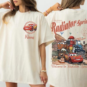 Op de afbeelding: Cr&egrave;mekleurig T-shirt met een rode raceauto-afbeelding en de naam "Name". De achterkant van het shirt heeft een vintage ontwerp met de tekst "Radiator Springs" en cartoon-autokarakters.