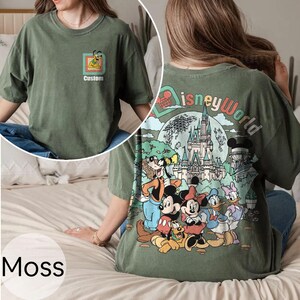 May include: Moss green t-shirt with a Disney World design on the back, showcasing Mickey Mouse, Minnie Mouse, Donald Duck, and friends. The front displays a smaller Pluto graphic and the word "Custom".