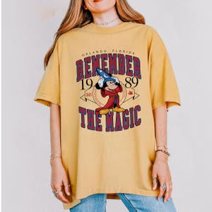 K&ouml;nnte beinhalten: Senfgelbes T-Shirt mit einer Grafik von Mickey Mouse in einem Zauberer-Outfit. Der Text auf dem Shirt lautet "REMEMBER 1989 THE MAGIC" mit "Orlando, Florida" &uuml;ber der Grafik. Das Shirt hat kurze &Auml;rmel und einen Rundhalsausschnitt.