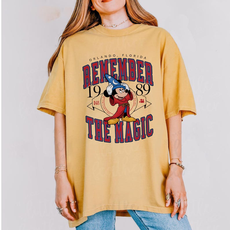 K&ouml;nnte beinhalten: Senfgelbes T-Shirt mit einer Grafik von Mickey Mouse in einem Zauberer-Outfit. Der Text auf dem Shirt lautet "REMEMBER 1989 THE MAGIC" mit "Orlando, Florida" &uuml;ber der Grafik. Das Shirt hat kurze &Auml;rmel und einen Rundhalsausschnitt.