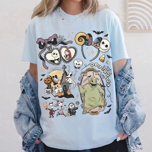 May include: A light blue t-shirt featuring a graphic print of characters and icons from The Nightmare Before Christmas, including Jack Skellington, Sally, Oogie Boogie, Zero, and Lock, Shock, and Barrel, arranged in a collage with bats and stars.