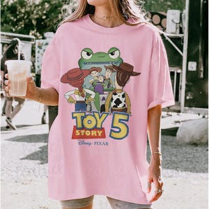 May include: Pink t-shirt featuring a graphic of characters from the movie Toy Story 5. The design includes Woody, Buzz Lightyear, and other characters. The shirt also has the Disney Pixar logo.