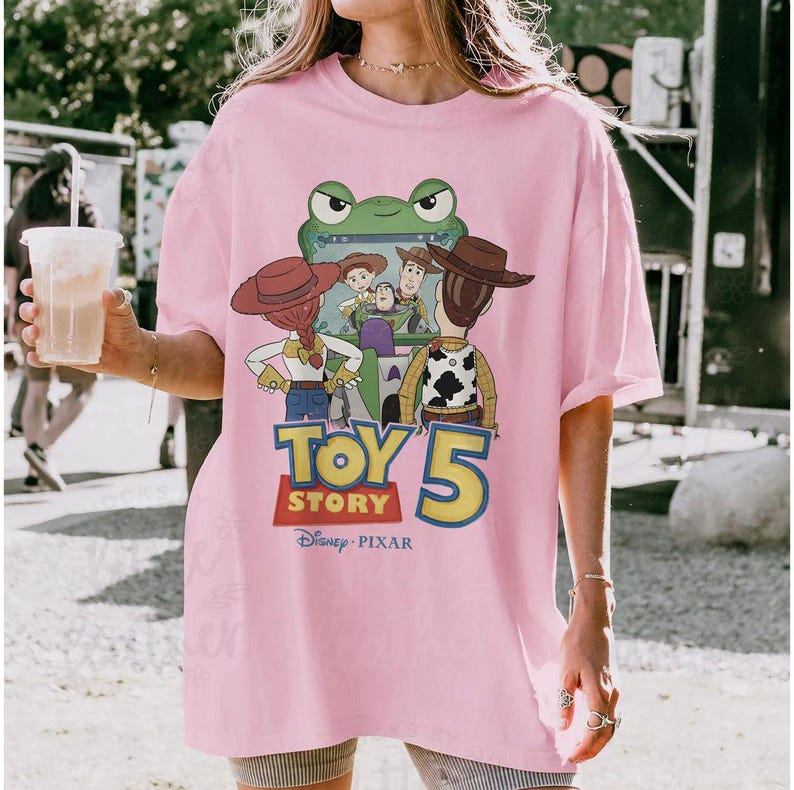 May include: Pink t-shirt featuring a graphic of characters from the movie Toy Story 5. The design includes Woody, Buzz Lightyear, and other characters. The shirt also has the Disney Pixar logo.
