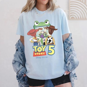 May include: Light blue t-shirt featuring a graphic of characters from the Toy Story movie franchise. The graphic includes a green frog, Woody, Buzz Lightyear, and the text "TOY STORY 5" in yellow and red. The Disney Pixar logo is at the bottom.
