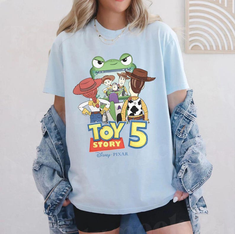 May include: Light blue t-shirt featuring a graphic of characters from the Toy Story movie franchise. The graphic includes a green frog, Woody, Buzz Lightyear, and the text "TOY STORY 5" in yellow and red. The Disney Pixar logo is at the bottom.