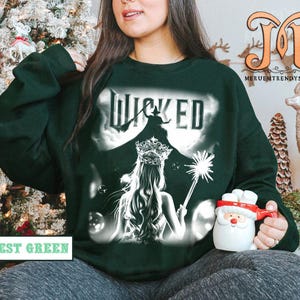 May include: Forest green sweatshirt with the word "Wicked" in white, featuring a silhouette of a witch and a woman with a crown. The sweatshirt has a graphic design and is a crew neck style.