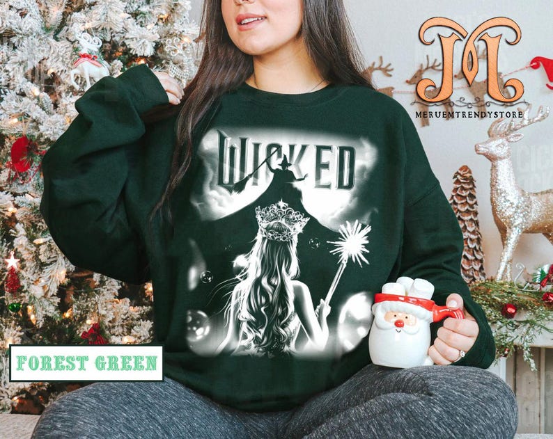 May include: Forest green sweatshirt with the word "Wicked" in white, featuring a silhouette of a witch and a woman with a crown. The sweatshirt has a graphic design and is a crew neck style.