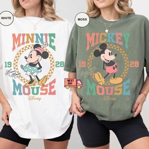 May include: Two t-shirts featuring vintage-style illustrations of Mickey and Minnie Mouse. The Mickey Mouse shirt is moss green with the text "Mickey Mouse" in rainbow colors. The Minnie Mouse shirt is white with the text "Minnie Mouse" in rainbow colors.