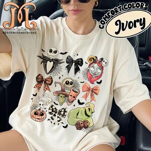Puede incluir: Camiseta de color marfil con personajes tem&aacute;ticos de Halloween y dise&ntilde;os de lazos. El gr&aacute;fico incluye a Jack Skellington, Sally, Oogie Boogie y otros personajes de The Nightmare Before Christmas. La camiseta tambi&eacute;n tiene el texto "COMFORT COLORS" e "Ivory".