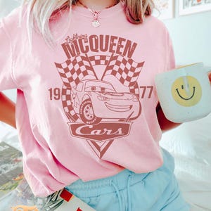 May include: Pink t-shirt with a vintage-style graphic of Lightning McQueen from the Disney Pixar movie Cars. The graphic features the text "Lightning McQueen" and "Cars" with a checkered flag background. The numbers 19 and 77 are also included in the graphic.