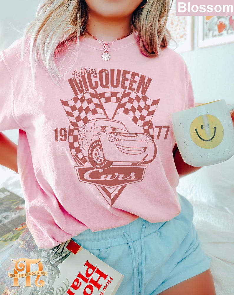 May include: Pink t-shirt with a vintage-style graphic of Lightning McQueen from the Disney Pixar movie Cars. The graphic features the text "Lightning McQueen" and "Cars" with a checkered flag background. The numbers 19 and 77 are also included in the graphic.
